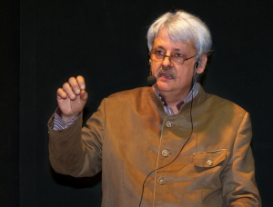 Ein älterer Mann mit grauem Haar und Brille, der eine braune Jacke mit Knöpfen und ein Headset-Mikrofon trägt, gestikuliert mit seiner rechten Hand, während er vor einem dunklen Hintergrund spricht.