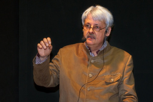 Ein älterer Mann mit grauem Haar und Brille, der eine braune Jacke mit Knöpfen und ein Headset-Mikrofon trägt, gestikuliert mit seiner rechten Hand, während er vor einem dunklen Hintergrund spricht.