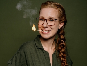 Eine lächelnde Frau mit Brille und langem Zopf hat ein brennendes Streichholz im Mund, aus dem eine kleine Flamme und Rauch aufsteigen, vor einem schlichten olivgrünen Hintergrund.