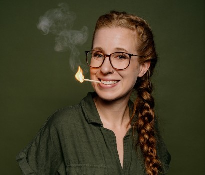 Eine lächelnde Frau mit Brille und langem Zopf hat ein brennendes Streichholz im Mund, aus dem eine kleine Flamme und Rauch aufsteigen, vor einem schlichten olivgrünen Hintergrund.