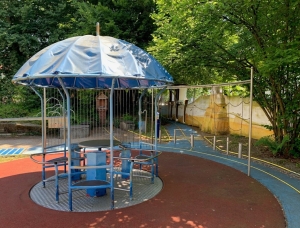 Ein Spielplatzbereich verfügt über eine blaue, schirmförmige Kletterstruktur mit Metallstangen und Sitzen darunter, umgeben von Gummiböden, Bäumen und einer gelben Wand im Hintergrund.