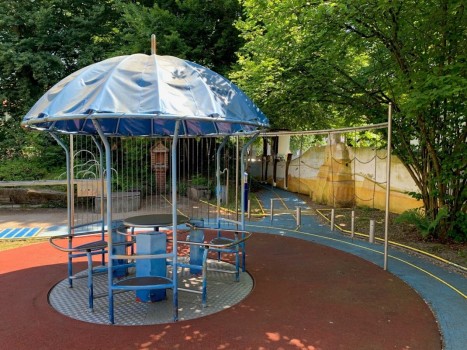 Ein Spielplatzbereich verfügt über eine blaue, schirmförmige Kletterstruktur mit Metallstangen und Sitzen darunter, umgeben von Gummiböden, Bäumen und einer gelben Wand im Hintergrund.