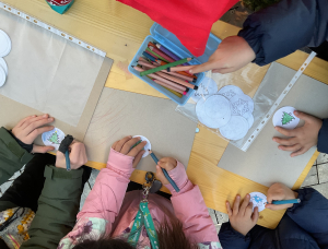 Kinder sitzen um einen Tisch und malen mit Buntstiften Weihnachtsschmuck aus Papier. Man sieht ihre Hände, wenn sie festliche Motive wie Bäume und Schneeflocken auf runde Ausschnitte zeichnen.