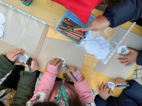 Kinder sitzen um einen Tisch und malen mit Buntstiften Weihnachtsschmuck aus Papier. Man sieht ihre Hände, wenn sie festliche Motive wie Bäume und Schneeflocken auf runde Ausschnitte zeichnen.