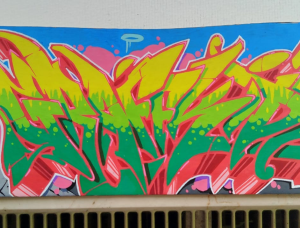 Buntes Graffiti-Wandbild mit einer Cartoon-Figur mit Mütze und Sonnenbrille auf der linken Seite, fetten gelben und grünen stilisierten Buchstaben in der Mitte und städtischen Gebäuden im Hintergrund vor einem blauen Himmel.