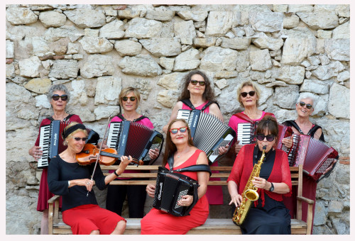 Sieben Frauen in roten und schwarzen Kleidern spielen Musikinstrumente, fünf Akkordeons, eine Geige und ein Saxophon, während sie vor einer Steinmauer posieren.