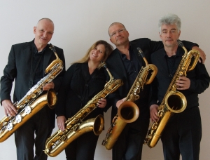 Vier schwarz gekleidete Musiker stehen nebeneinander, jeder hält ein Saxophon, lächelt und posieren vor einem schlichten, hellen Hintergrund.