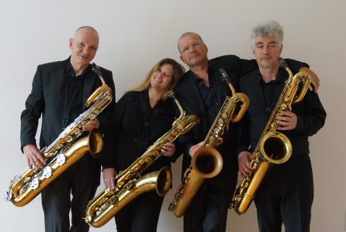 Vier schwarz gekleidete Musiker stehen nebeneinander, jeder hält ein Saxophon, lächelt und posieren vor einem schlichten, hellen Hintergrund.