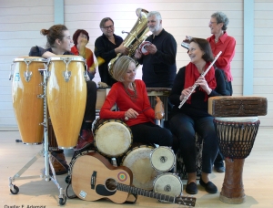 Eine Gruppe von sieben Personen mit verschiedenen Musikinstrumenten, darunter Congas, eine Tuba, Trommeln, eine Gitarre, eine Flöte und eine Djembe, sitzen und stehen zusammen in einem Innenbereich, lächeln und interagieren miteinander.