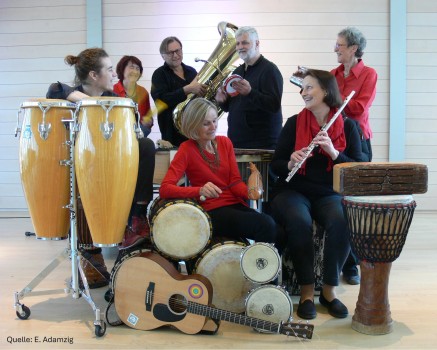 Eine Gruppe von sieben Personen mit verschiedenen Musikinstrumenten, darunter Congas, eine Tuba, Trommeln, eine Gitarre, eine Flöte und eine Djembe, sitzen und stehen zusammen in einem Innenbereich, lächeln und interagieren miteinander.