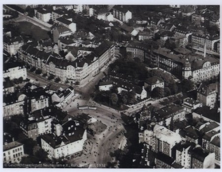 Eine Schwarz-Weiß-Luftaufnahme einer Stadtkreuzung mit mehreren zusammenlaufenden Straßen, umgeben von historischen mehrstöckigen Gebäuden, Grünflächen und Bäumen. Die Szene scheint aus dem frühen 20. Jahrhundert zu stammen.
