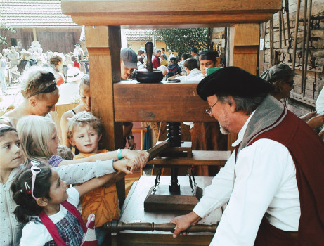 Kinder versammeln sich um eine historische Druckerpresse und beobachten, wie ein älterer Mann in einem historischen Kostüm die Maschine bedient. Viele Menschen sind anwesend.