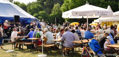 Menschen sitzen an Holztischen unter großen weißen Sonnenschirmen und genießen Essen und Getränke auf einem lebhaften Fest oder Jahrmarkt. Im Hintergrund spielt eine Band unter einem blau-weißen Zelt auf der Bühne. Bäume und Sonnenschein runden die Szene ab.