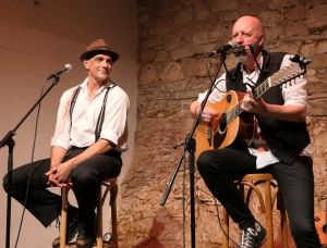 Zwei Männer sitzen auf Hockern auf der Bühne. Der eine, mit Hut und Hosenträgern, lächelt, während der andere, in eine Weste gekleidet, in ein Mikrofon singt und eine akustische Gitarre spielt. Im Hintergrund ist eine Steinmauer zu sehen.
