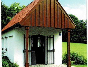 Ansicht der Echadinger Kapelle mit rotem Giebeldach und Holzbalken, mit kopfsteingepflastertem Weg im Vordergrund, umgeben von grünem Gras und Bäumen unter einem klaren blauen Himmel.
