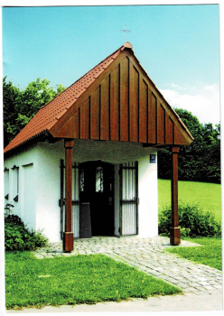 Ansicht der Echadinger Kapelle mit rotem Giebeldach und Holzbalken, mit kopfsteingepflastertem Weg im Vordergrund, umgeben von grünem Gras und Bäumen unter einem klaren blauen Himmel.