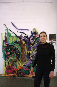 Eine Frau in Schwarz steht vor einer farbenfrohen Textilkunstinstallation an einer Wand, die abstrakte Formen in Grün, Braun, Lila und Rosa zeigt. An der rechten Seite der Wand ist ein kleines Foto angeheftet.