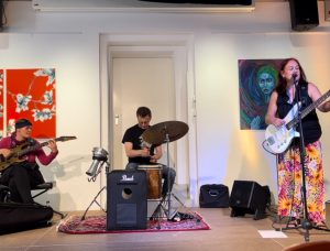 eine Musikerin mit Gitarre und zwei Musiker mit Schlagzeug und Bass spielen in einem Saal