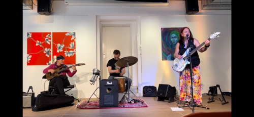 eine Musikerin mit Gitarre und zwei Musiker mit Schlagzeug und Bass spielen in einem Saal