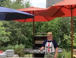 Ein Mann mit weißen Bart steht vor eiem Gasgrill unter drei Sonnenschirmen. Er hält dabei seine Grillzange in der rechten Hand