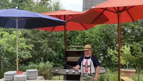 Ein Mann mit weißen Bart steht vor eiem Gasgrill unter drei Sonnenschirmen. Er hält dabei seine Grillzange in der rechten Hand