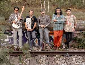 Fünf Männer stehen an einer mit Graffiti bedeckten Wand in der Nähe von Bahngleisen. Zwei halten Instrumente – eine weiße E-Gitarre und ein Keyboard –, ein dritter hat einen Bass zu seinen Füßen. Sie posieren im Grünen und blicken in die Kamera.