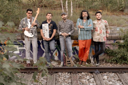Fünf Männer stehen an einer mit Graffiti bedeckten Wand in der Nähe von Bahngleisen. Zwei halten Instrumente – eine weiße E-Gitarre und ein Keyboard –, ein dritter hat einen Bass zu seinen Füßen. Sie posieren im Grünen und blicken in die Kamera.