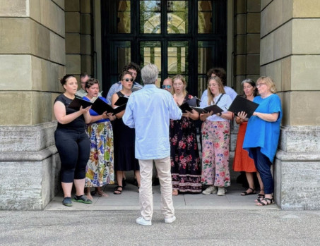 Eine Gruppe von Menschen steht in einem Torbogen und singt aus schwarzen Ordnern, während eine Person in einem hellblauen Hemd ihnen gegenübersteht und dirigiert. Die Sänger sind leger gekleidet und stehen vor einem Steingebäude mit hohen Türen.