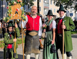 Vier Personen in traditioneller bayerischer Tracht stehen im Freien und lächeln. Drei Erwachsene und ein Kind tragen Hüte, Westen und Lederhosen oder Dirndl. Das Kind hält ein dekoratives Schild, die Erwachsenen halten Stäbe. Im Hintergrund sind Gebäude und Bäume zu sehen.