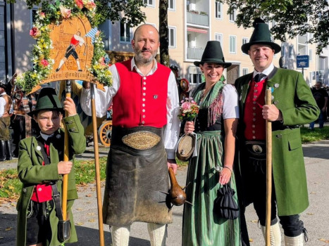 Vier Personen in traditioneller bayerischer Tracht stehen im Freien und lächeln. Drei Erwachsene und ein Kind tragen Hüte, Westen und Lederhosen oder Dirndl. Das Kind hält ein dekoratives Schild, die Erwachsenen halten Stäbe. Im Hintergrund sind Gebäude und Bäume zu sehen.