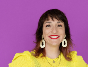 Eine Person mit schulterlangen braunen Haaren, die ein leuchtend gelbes Oberteil, eine blaue Hose, große weiße Ohrringe und roten Lippenstift trägt, steht lächelnd und mit den Händen in den Hüften vor einem lila Hintergrund.