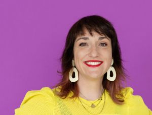 Eine Person mit schulterlangen braunen Haaren, die ein leuchtend gelbes Oberteil, eine blaue Hose, große weiße Ohrringe und roten Lippenstift trägt, steht lächelnd und mit den Händen in den Hüften vor einem lila Hintergrund.