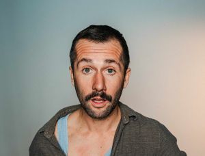Ein Mann mit Schnurrbart hält ein Mikrofon in der Hand und zieht sein Hemd auf, so dass ein blaues Unterhemd zum Vorschein kommt. Er sieht überrascht aus und steht vor einem schlichten Hintergrund.