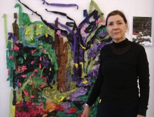 Eine Frau in Schwarz steht vor einer farbenfrohen Textilkunstinstallation an einer Wand, die abstrakte Formen in Grün, Braun, Lila und Rosa zeigt. An der rechten Seite der Wand ist ein kleines Foto angeheftet.