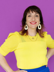 Eine Person mit schulterlangen braunen Haaren, die ein leuchtend gelbes Oberteil, eine blaue Hose, große weiße Ohrringe und roten Lippenstift trägt, steht lächelnd und mit den Händen in den Hüften vor einem lila Hintergrund.