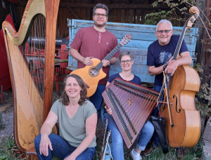 Vier lächelnde Menschen posieren im Freien mit Musikinstrumenten: einer Harfe, einer Gitarre, einem Hackbrett und einem Kontrabass. Sie sitzen und stehen vor einer rustikalen blauen Holzbank und einer Scheunenkulisse.