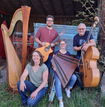Vier lächelnde Menschen posieren im Freien mit Musikinstrumenten: einer Harfe, einer Gitarre, einem Hackbrett und einem Kontrabass. Sie sitzen und stehen vor einer rustikalen blauen Holzbank und einer Scheunenkulisse.