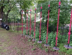 Ein Hindernisparcours auf einem Spielplatz mit roten Seilschaukeln und in einer Reihe hängenden Holzplattformen, neben schwarzen Reifenschaukeln und weißen Kletterseilen, umgeben von grünen Bäumen und Büschen.