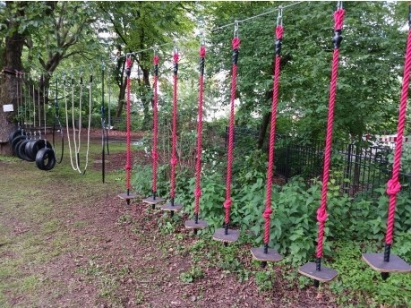 Ein Hindernisparcours auf einem Spielplatz mit roten Seilschaukeln und in einer Reihe hängenden Holzplattformen, neben schwarzen Reifenschaukeln und weißen Kletterseilen, umgeben von grünen Bäumen und Büschen.