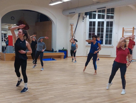 Eine Gruppe von Personen nimmt an einem Fitness- oder Tanzkurs in einer geräumigen Halle teil und folgt einer weiblichen Lehrerin, die sich mit erhobenem Arm energisch bewegt.