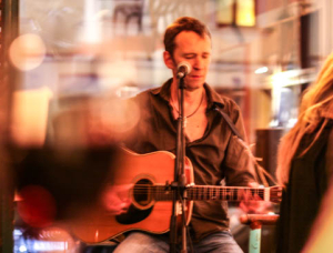 Ecco Meineke spielt eine Akustikgitarre und singt in ein Mikrofon in einer Halle, mit warmer Beleuchtung und unscharfem Vordergrund.