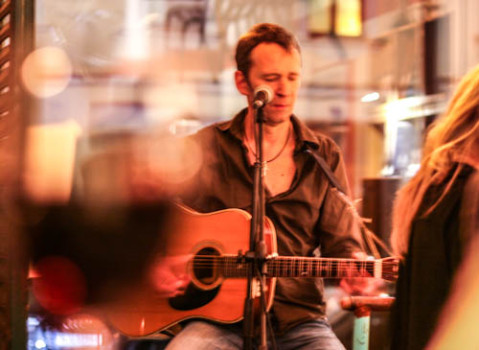 Ecco Meineke spielt eine Akustikgitarre und singt in ein Mikrofon in einer Halle, mit warmer Beleuchtung und unscharfem Vordergrund.