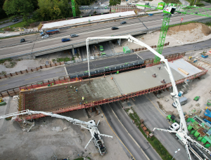 Luftaufnahme der im Bau befindlichen Autobahnüberführung Kreuzhof mit zwei großen Betonpumpenfahrzeugen, die Beton auf das Bauwerk gießen; Arbeiter und Baumaschinen sind zu sehen. In der Nähe befindliche Straßen und Fahrzeuge sind zu sehen.