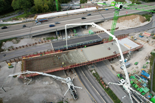 Luftaufnahme der im Bau befindlichen Autobahnüberführung Kreuzhof mit zwei großen Betonpumpenfahrzeugen, die Beton auf das Bauwerk gießen; Arbeiter und Baumaschinen sind zu sehen. In der Nähe befindliche Straßen und Fahrzeuge sind zu sehen.