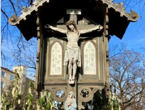 Berg-am-Laimer Kreuzweg-Marterl: Ein hölzernes Kruzifix im Freien mit einer Statue von Jesus am Kreuz unter einem kleinen Dach und einer Figur von Maria darunter, umgeben von Bäumen und Grün, mit blauem Himmel und Gebäuden im Hintergrund.