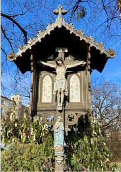 Berg-am-Laimer Kreuzweg-Marterl: Ein hölzernes Kruzifix im Freien mit einer Statue von Jesus am Kreuz unter einem kleinen Dach und einer Figur von Maria darunter, umgeben von Bäumen und Grün, mit blauem Himmel und Gebäuden im Hintergrund.