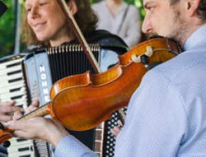 Eine Frau, die Akkordeon spielt, und ein Mann, der Geige spielt, musizieren zusammen in einem Haus, beide konzentriert und lächelnd. Eine verschwommene Person sitzt im Hintergrund.