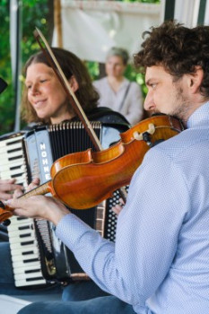 Eine Frau, die Akkordeon spielt, und ein Mann, der Geige spielt, musizieren zusammen in einem Haus, beide konzentriert und lächelnd. Eine verschwommene Person sitzt im Hintergrund.
