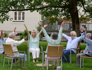 Sechs ältere Menschen sitzen im Freien im Kreis unter einem Baum und heben während einer Gruppenübung oder Entspannungssitzung die Arme. Sie sitzen auf Stühlen und scheinen sich in einem Garten in der Nähe eines Gebäudes zu befinden.