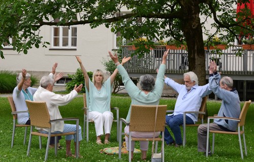 Sechs ältere Menschen sitzen im Freien im Kreis unter einem Baum und heben während einer Gruppenübung oder Entspannungssitzung die Arme. Sie sitzen auf Stühlen und scheinen sich in einem Garten in der Nähe eines Gebäudes zu befinden.